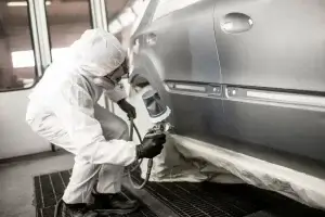 A person in protective gear is painting a car in a workshop using a spray gun.