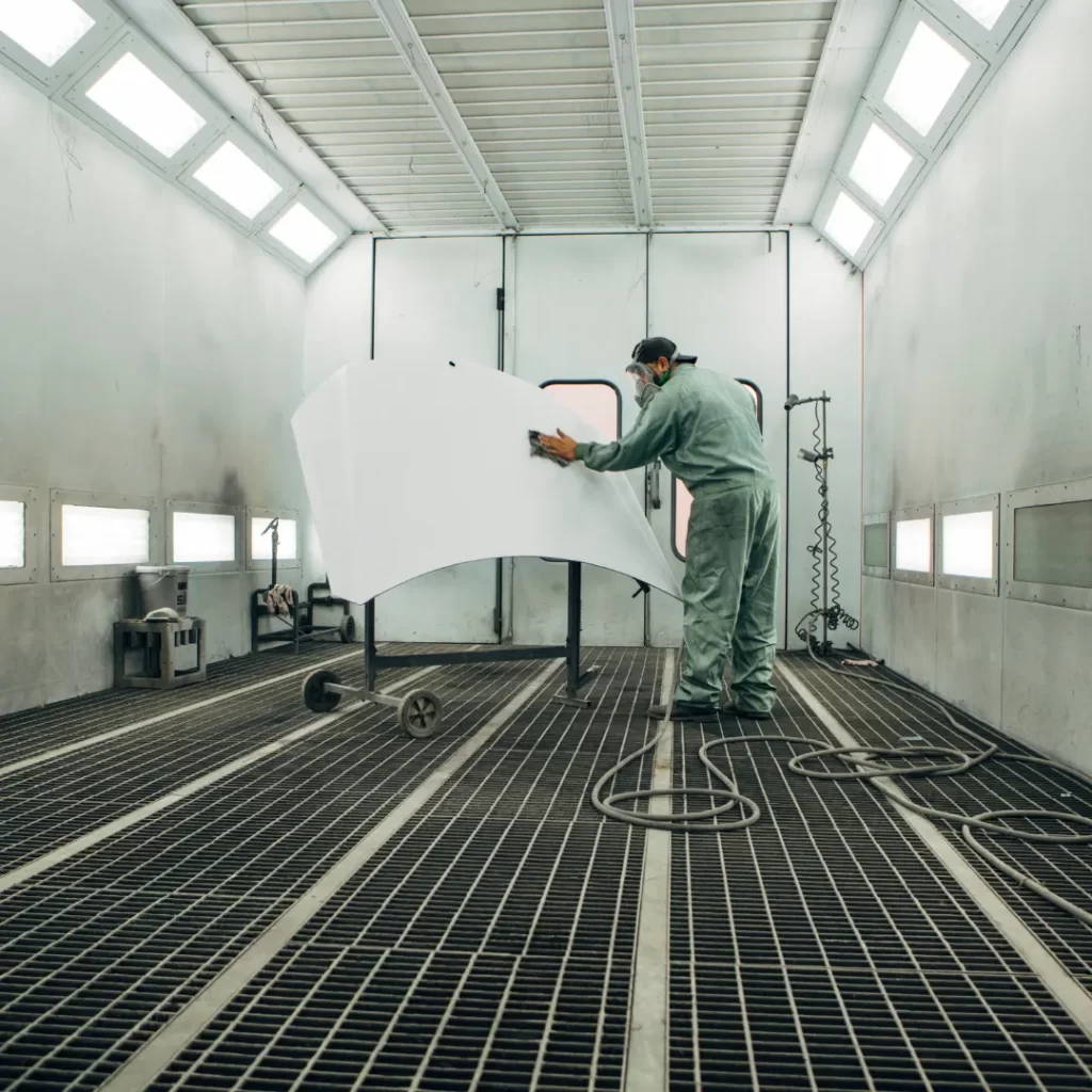 A worker in protective gear sands a large white vehicle panel in a well-lit industrial spray paint booth.